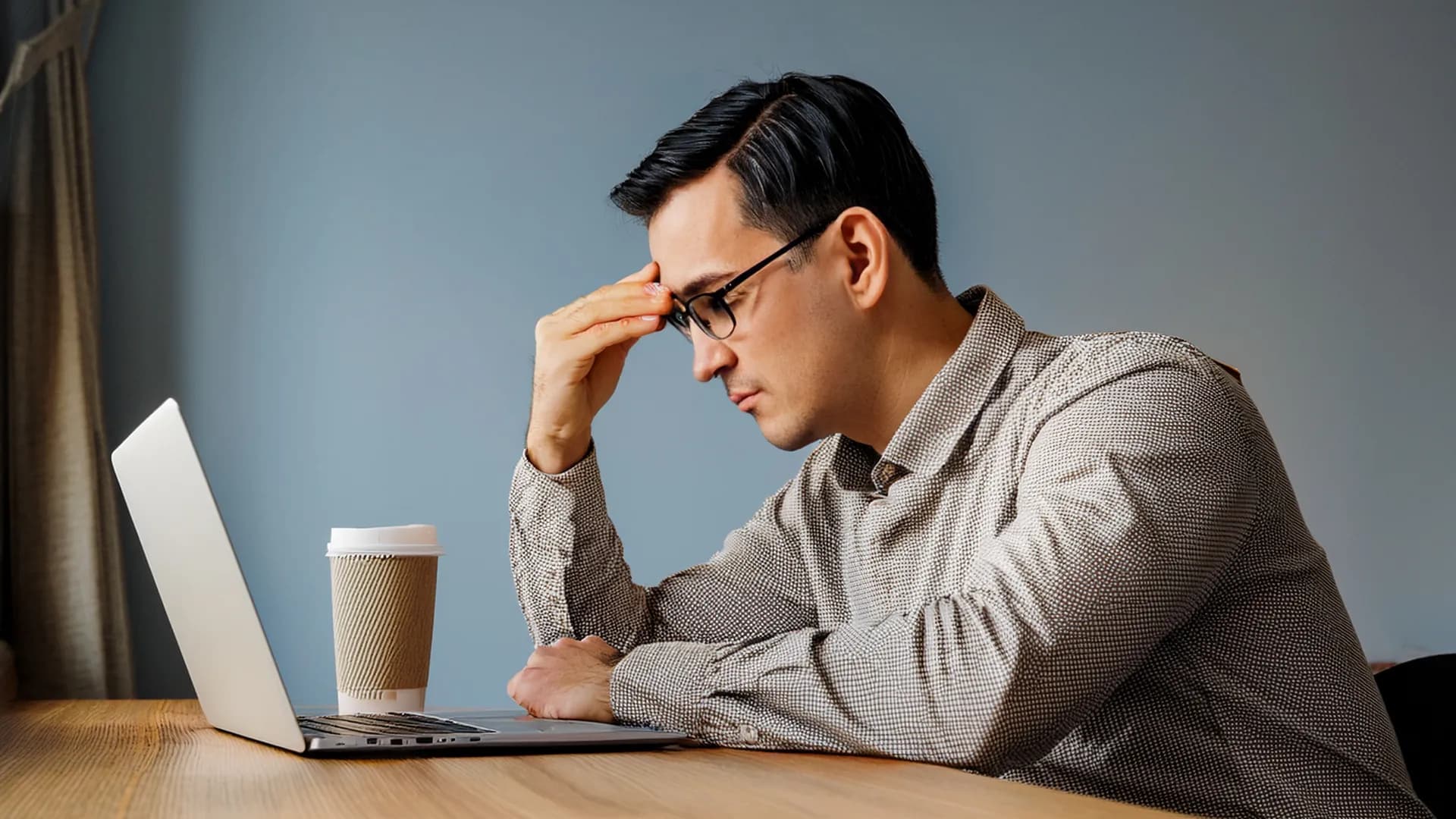 A person at a desk leaning forward with one hand on their forehead, staring at a laptop screen with a frustrated expression, warm natural daylight from the right